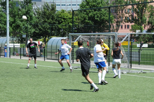 Leute in Kleidung beim Fussballspielen auf einem Feld mit einem Zaun und einem Tor in der Mitte, einen Pfahl und einen Fussball oben links und Bäume und Gebäude im Hintergrund.
