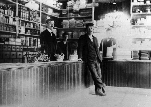 Schwarz-weiß-Foto von drei Männern an einer Ladentheke mit Gegenständen, einem Hintergrundgestell mit Büchern und Artikeln und einer "The New York Times"-überschrift, die einen Raub suggeriert.