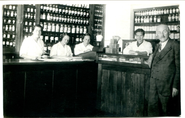 Ein Schwarz-Weiß-Foto einer Gruppe von Menschen, die vor einer Theke in einer Apotheke stehen, mit Papieren und Gegenständen auf der Theke und Regalen voller Flaschen im Hintergrund.