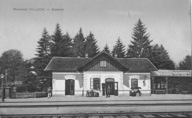 Ein altes Schwarz-Weiß-Foto eines Bahnhofs in Warmbad Villach, Bahnhof, mit einem Haus mit Fenstern und Türen, einer Gruppe von Menschen, die auf Bänken stehen und sitzen, einem Zaun, einem Straßenpfahl, einer Schautafel, einigen Bäumen und einem bewölkten Himmel.