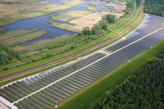 Luftaufnahme einer Solar-Farm mit Panelen in einem Feld, umgeben von Bäumen, Gras, Pflanzen und Wasser, mit einem Zug auf einer nahen Eisenbahnschiene.