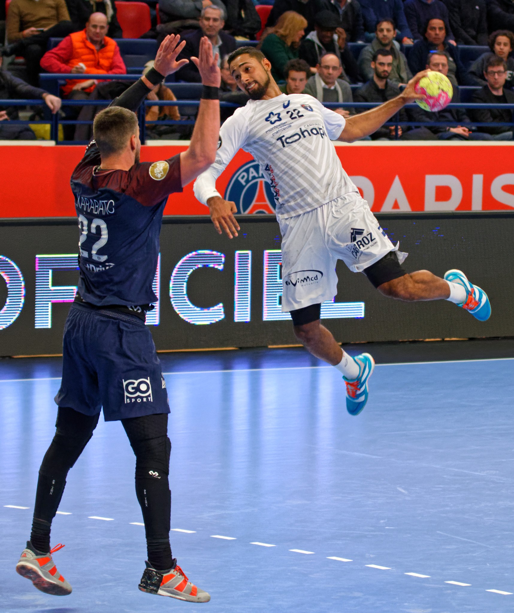 Zwei Männer bei einem Handballspiel auf einem Platz, einer springt in der Luft und hält den Ball, mit Zuschauern und einer Anzeigetafel im Hintergrund.
