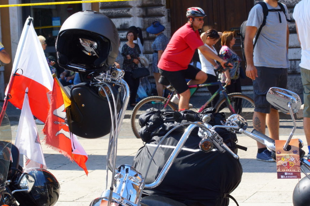 Mehrere Motorräder mit Helmen und Fahnen sowie eine schwarze Tasche und Personen im Hintergrund, eine sitzend auf einem Motorrad mit Sonnenbrille und Helm.