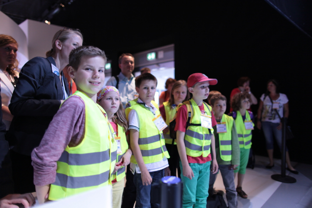 Eine Gruppe von Kindern in Sicherheitswesten und Namensschildern auf einem Boden stehend, mit einem Jungen auf der linken Seite, der lächelt, einem Pfahl auf der rechten Seite und einigen Menschen im Hintergrund.