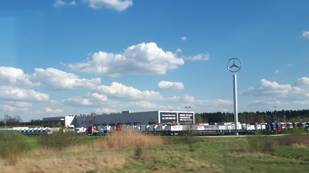 Ein Mercedes-Benz-Werk in Deutschland mit Fahrzeugen auf der Straße, Grünflächen, Gebäuden und einem Logo-Pfahl unter einem bewölkten Himmel.