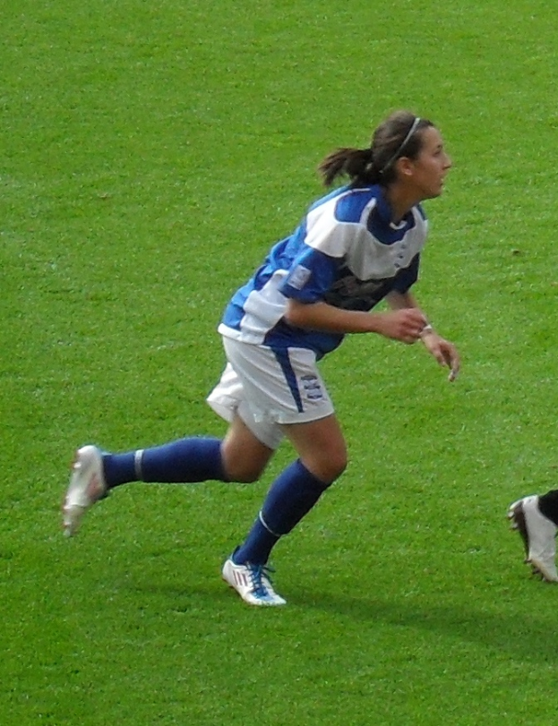 Eine Frau in einem blauen und weißen Trikot läuft auf einem Fußballfeld, wobei ein Bein einer Person auf der rechten Seite des Bildes zu sehen ist.