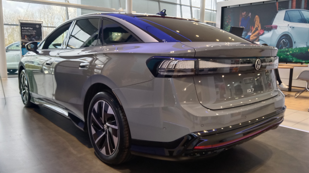 Ein Volkswagen ID 7 Elektroauto in einer Ausstellungshalle, mit einer Person auf der rechten Seite, anderen Autos, einer Tafel und Glaswänden im Hintergrund.