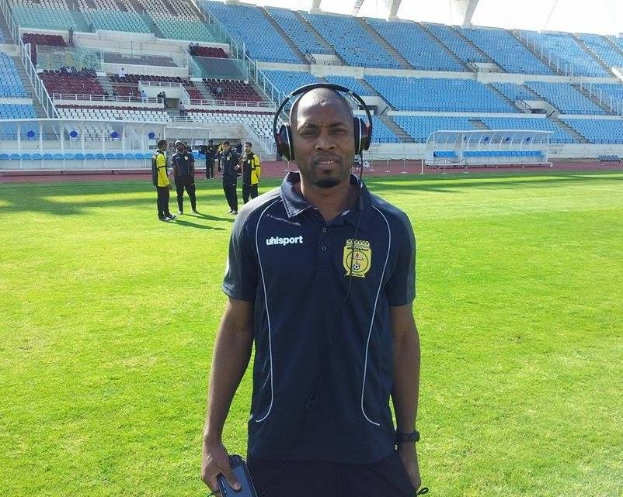 Ein Mann in einem Headset, wahrscheinlich der Trainer der ghanaischen Nationalmannschaft, steht auf einem Fussballfeld in der Nähe eines Stadions und hält ein Objekt in der Hand, während im Hintergrund Menschen zu sehen sind.
