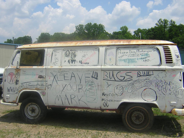 Ein beschädigtes Wohnmobil mit Gemälden und Zitaten darauf, das vor einem bewaldeten Gebiet geparkt ist.