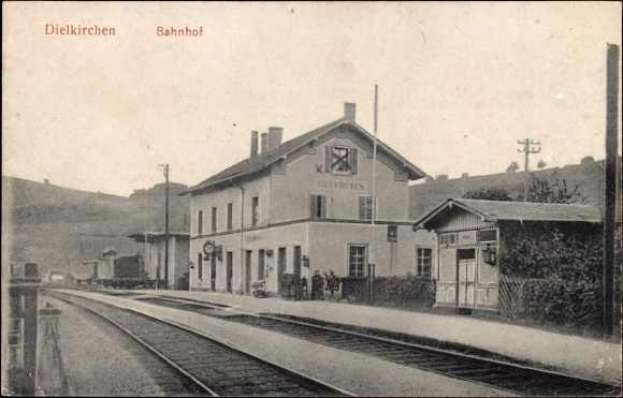 Schwarzes und weißes Foto eines Bahnhofs in Dielkirchen, Deutschland, mit einem Zug auf den Gleisen, umgeben von Häusern, Pfählen, Bäumen, Pflanzen und Menschen, Hügel im Hintergrund und einem klaren Himmel.
