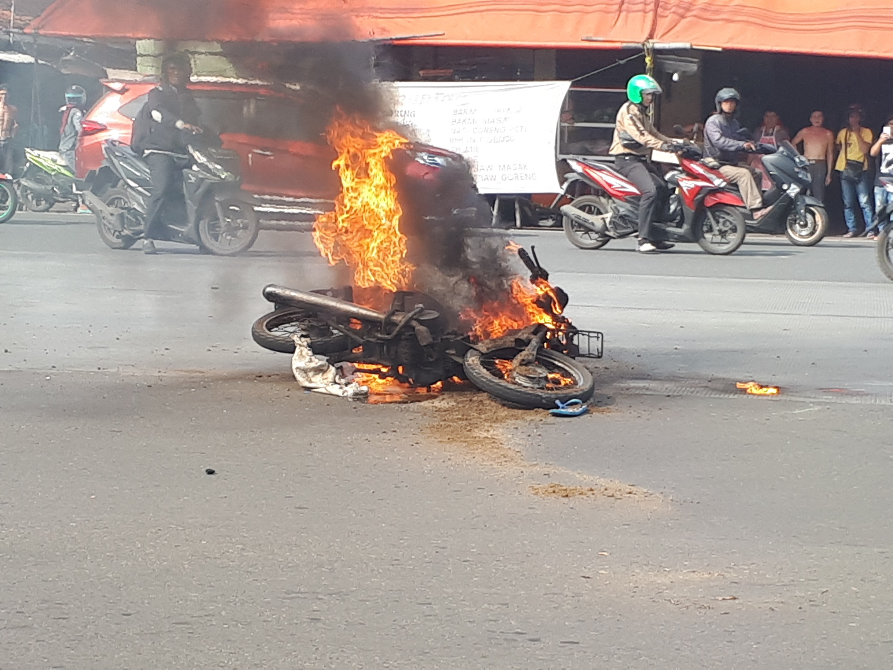 Motorrad in Flammen auf der Straße mit einem Auto und einem Van im Hintergrund, umgeben von Menschen, einige tragen Helme, mit einem Banner und einem Zelt im Bild.