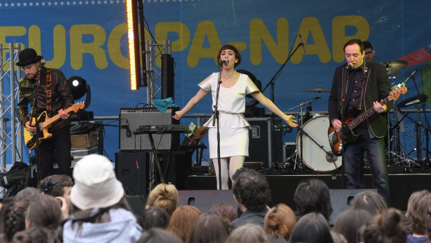 Vier Personen auf der Bühne mit einer Frau in einem weißen Kleid, die singt, und zwei Männern, die Gitarren spielen, während eine Menge vor Lautsprechern und einer Wand zusieht.