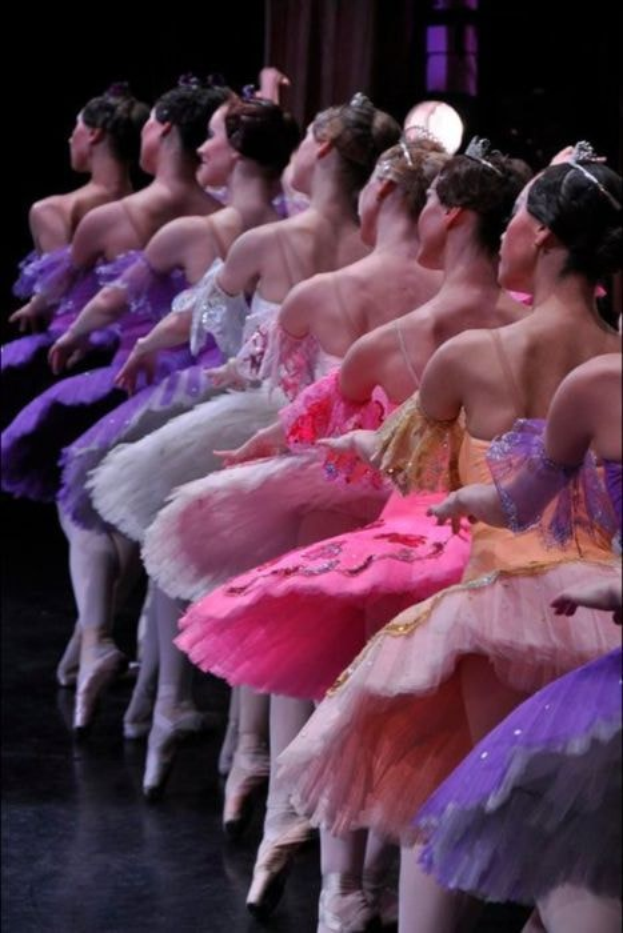 Eine Gruppe von Balletttänzerinnen in rosafarbenen Tutus sitzt auf einer Bühne, beleuchtet von einem Licht im Hintergrund, und bewegt sich in perfekter Harmonie.