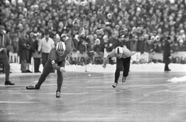 Zwei Männer im Eisschnelllauf auf einer Eisbahn vor einem großen Publikum, mit Helmen und Eislaufschuhen, mit einem unscharfen Hintergrund, in Schwarz-Weiß.