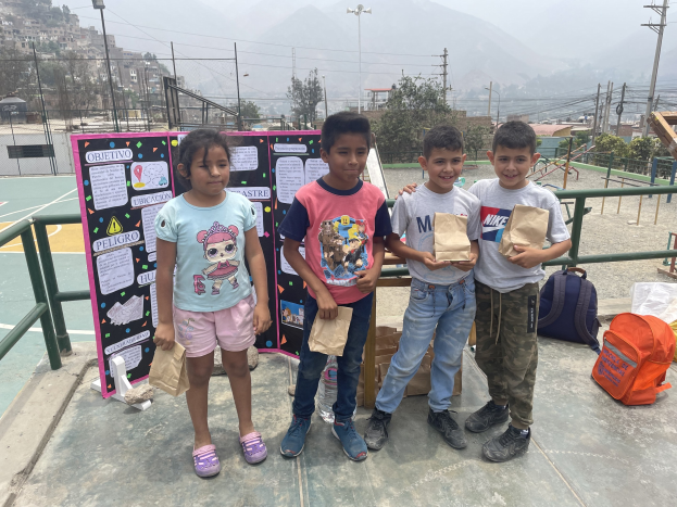 Gruppe von Kindern auf einem Basketballfeld mit Papiertüten, mit Vorräten auf dem Boden, eine Tafel mit Text links und ein Hintergrund aus Zaun, Pfosten, Straßenlaternen, Bäumen, Gebäuden, Hügeln und Himmel.