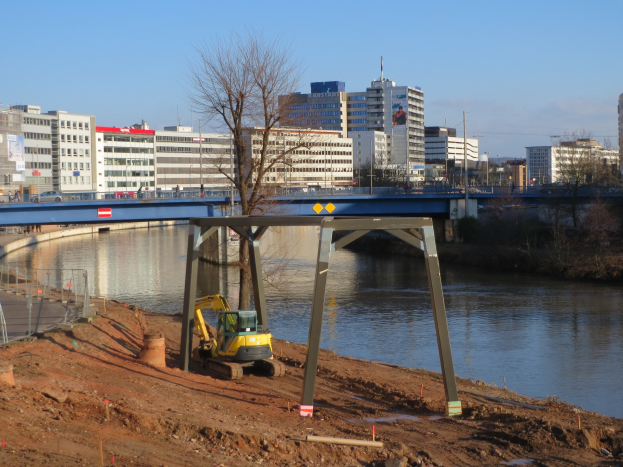 Eine Baustelle mit einer Brücke über einen Fluss, ein Bagger im Vordergrund, eine Straße mit einem Geländer auf der linken Seite und Bäume, Gebäude, Pfähle und einen klaren blauen Himmel im Hintergrund.