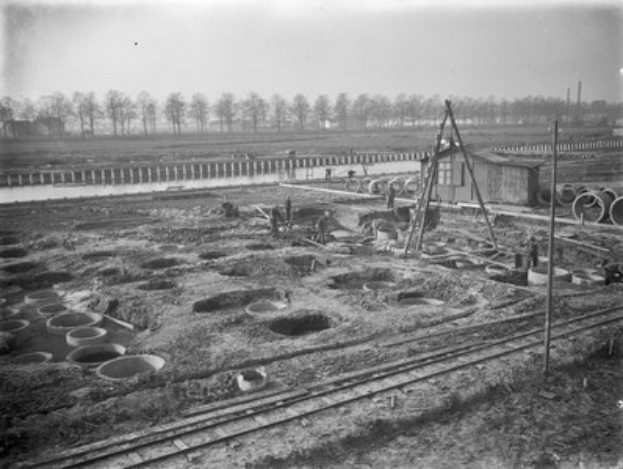 Schwarzes und weißes Foto einer Baustelle mit einer Bahnschiene, Menschen, Rohren, Pfählen, einem Gewässer, einer Brücke und Bäumen im Hintergrund.