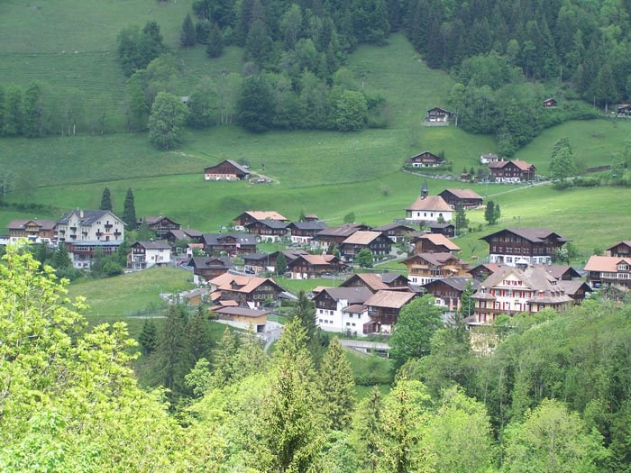 Kleines Dorf mit bunten Häusern in einer grünen Talsenke umgeben von Bäumen und Gras.
