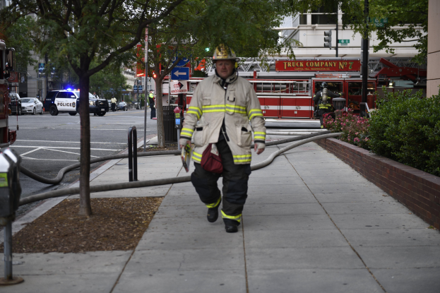 Feuerwehrmann geht auf einem Bürgersteig mit einem Schlauch in der Hand, trägt einen Helm und ist von städtischen Elementen und Vegetation umgeben, mit Gebäuden und Himmel im Hintergrund.