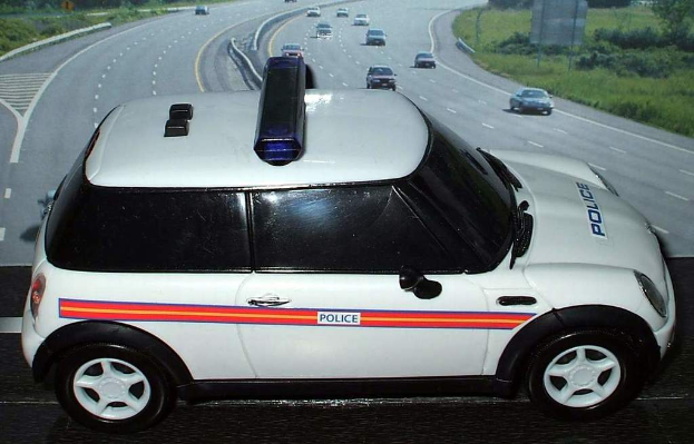 Ein weißes Polizeiauto ist im Vordergrund zu sehen, dahinter bewegen sich viele Autos auf der Straße, umgeben von Gras und Bäumen auf beiden Seiten.