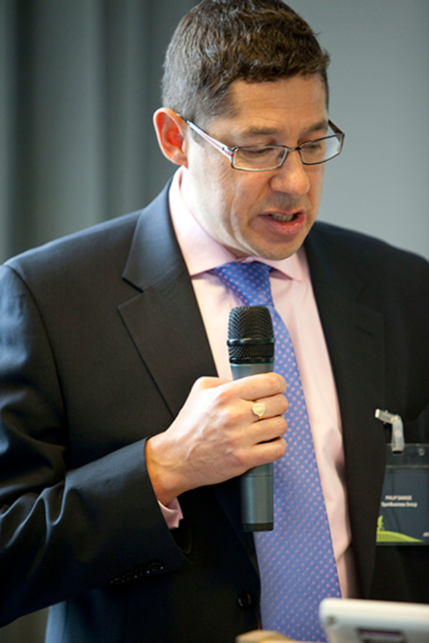 Ein Mann in Anzug, Hemd und Krawatte mit Brille hÃ¤lt ein Mikrofon und spricht, mit einem weiÃen Abzeichen an seinem Hemd und einem Vorhang im Hintergrund.