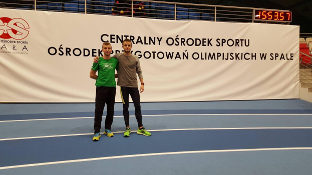 Zwei Männer stehen auf einem Tennisplatz mit einer Tafel im Hintergrund, auf der "Olympische Spiele 2019 - Ostrodek vs Spale" steht, sowie einer Digitalanzeigetafel, einem Geländer und ein paar Stühlen.