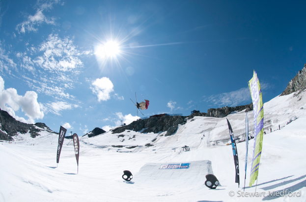 Eine Person in der Luft mit Skiern, mit schneebedeckten Bergen im Hintergrund.