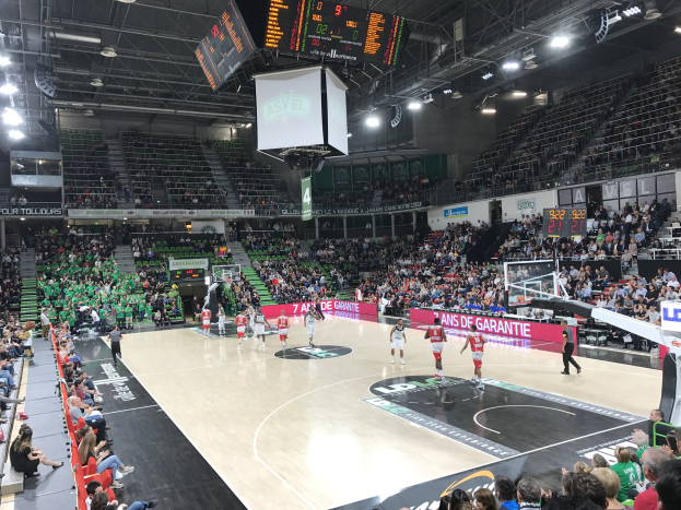 Basketball-Spiel zwischen Asvel und Galatasaray in einer großen Halle mit Zuschauern, Anzeigetafeln und Deckenbeleuchtung und -bildschirm.