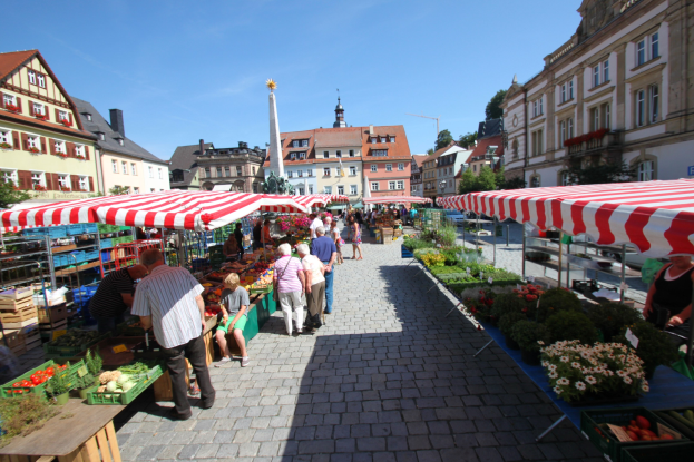 Ein belebter Markt im historischen Zentrum von Heidelberg mit Menschen, die gehen, auf Bänken sitzen und in der Nähe von Zelten stehen, mit Gemüsekörben auf Tischen, Gebäuden mit Fenstern, Bäumen und einem klaren blauen Himmel im Hintergrund.