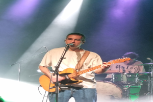 Ein Mann spielt eine E-Gitarre auf der Bühne, beleuchtet von Scheinwerfern von oben, während er in ein Mikrofon singt und ein anderer Mann Schlagzeug spielt im Hintergrund.