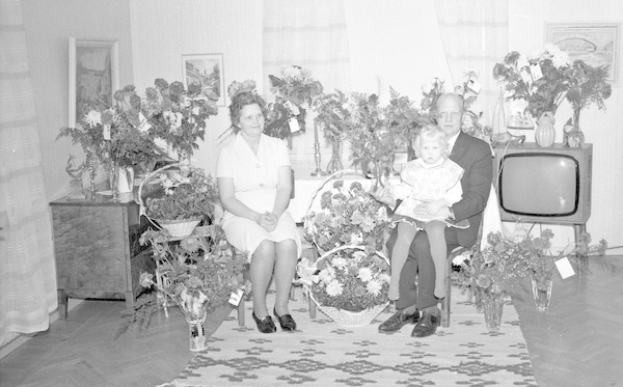 Schwarzes und weißes Foto einer Familie aus drei Personen - einem Mann, einer Frau und einem Kind - auf einem Sofa in einem Wohnzimmer mit Blumenvasen, Teppich, Vorhängen und Fotorahmen im Hintergrund.