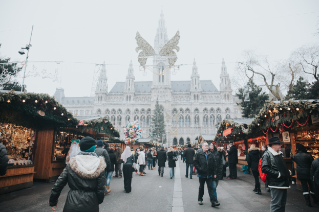 Ein belebter Weihnachtsmarkt in Wien, Österreich, mit Menschen, die herumlaufen, Ständen, die mit Lichtern und festlichen Artikeln geschmückt sind, einem Gebäude mit Fenstern im Hintergrund, Bäumen und einem klaren blauen Himmel.