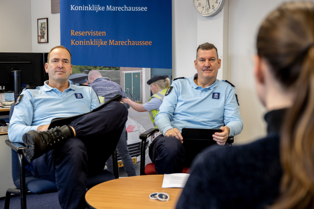 Zwei Polizeibeamte sitzen an einem Tisch mit einer Frau, Papieren und Gegenständen auf dem Tisch, einer Wand mit einem Banner, einer Uhr und Fotorahmen im Hintergrund und einem weiteren Tisch mit einem Monitor, Tastatur und Maus links daneben.
