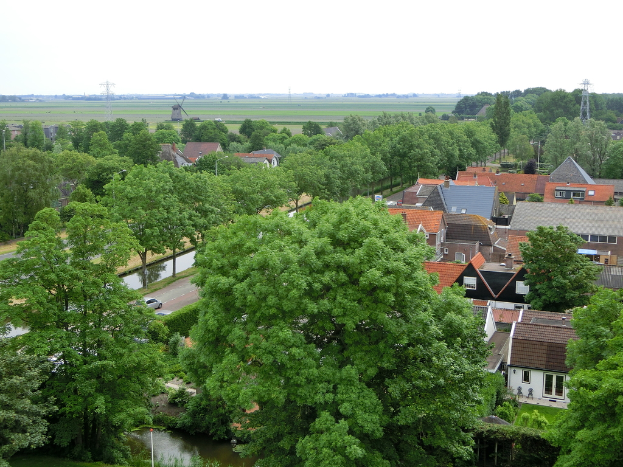 Eine Stadtrandszene mit H├Ąusern, B├Ąumen und Autos auf einer Stra├če, mit Turbinen und einer Windm├╝hle im Hintergrund.
