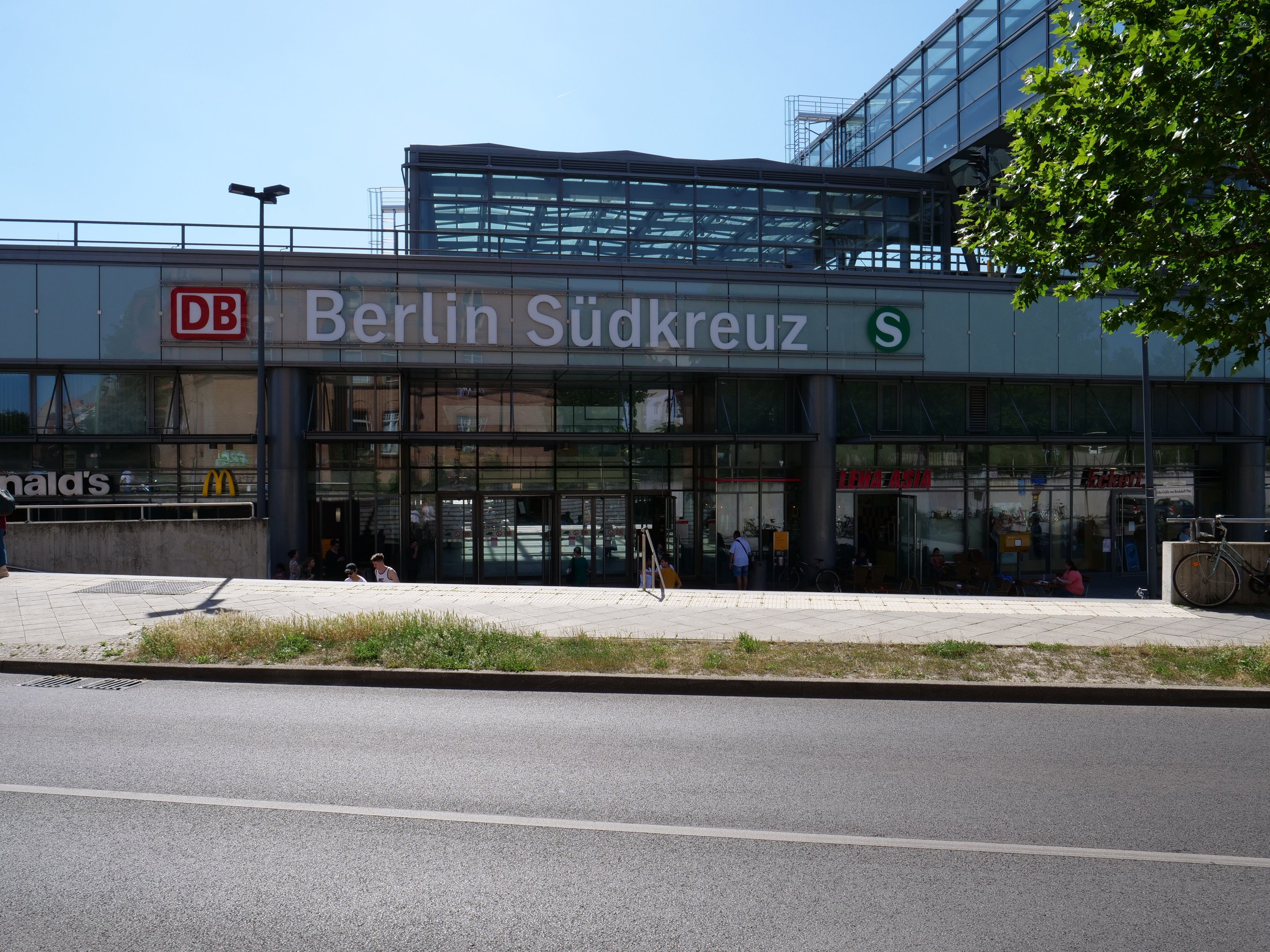 Ein gro├če Glaswand-Bahnhof namens "Berlin Südkreuz" in Berlin, Deutschland, mit umliegender Stra├čeninfrastruktur, Fahrzeugen, Fü├čgängern, Fahrr├Ądern, B├Ąumen und einem klaren blauen Himmel.