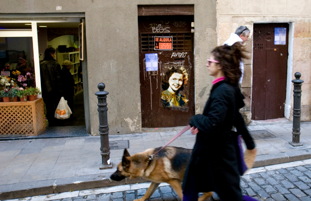Ein Mädchen spaziert mit ihrem Hund auf einer Straße, mit einem Geschäft, einigen Menschen und einem Mann auf dem Bürgersteig im Hintergrund und einer Wand, die mit Stickern und Plakaten bedeckt ist.