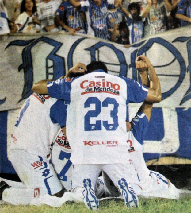 Gruppe von Fußballspielern, die vor jubelnder Menge auf dem Boden feiern, mit einem Banner im Hintergrund.