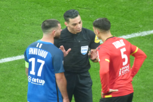 Ein Schiedsrichter in schwarzer Kleidung spricht mit zwei Männern auf einem Fötballfeld, einer in blauen und roten T-Shirts mit Text, beide tragen Brillen.