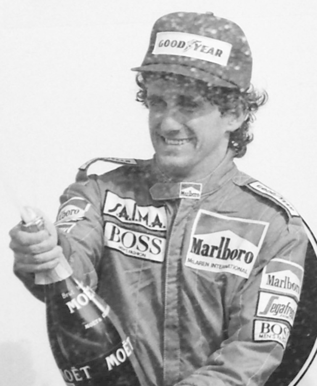 Ayrton Senna, ein Formel-Eins-Fahrer, steht lachend in einem Rennanzug und einer Mütze, hält eine Champagnerflasche in der Hand.