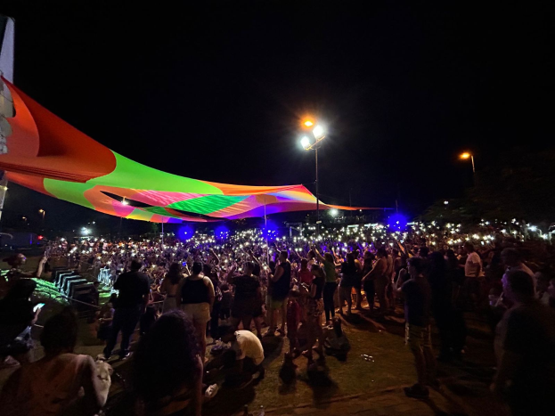 Eine große Menschenmenge steht vor einem Zelt im Dunkeln, beleuchtet von bunten Lichtern und Pfählen, mit einem Gebäude auf der linken Seite und dem Nachthimmel im Hintergrund während des Festivals der Künste im Jahr 2018.