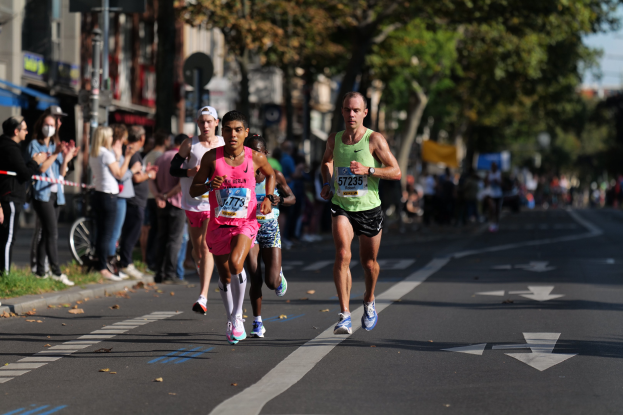 Gruppe von Menschen, die in einem Stadtmarathon laufen, mit Zuschauern auf der linken Seite, unscharfem städtischem Hintergrund mit Bäumen, Gebäuden und einem Fahrrad.
