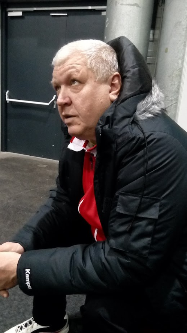 Ein Mann in einer schwarzen Jacke und einem roten Hemd sitzt auf dem Boden vor einer Tür mit einem Griff, einer Säule und einer Wand und sieht tief nachdenklich aus.