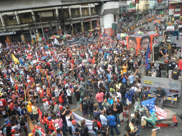 Eine große Menschenmenge geht eine Straße entlang und hält Fahnen, Schirme und Plakate hoch, während Autos, Verkehrszeichen, Gebäude, Schilder und eine Brücke im Hintergrund zu sehen sind.