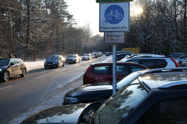 Ein Stau auf einer verschneiten Straße mit parkenden Autos am Straßenrand, ein Verkehrsschild im Vordergrund und Bäume mit einem klaren Himmel im Hintergrund.