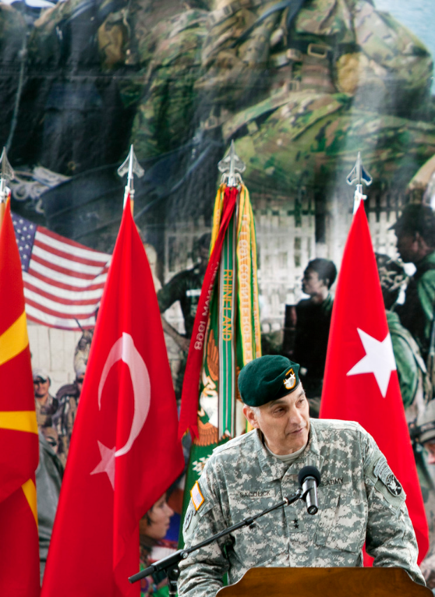 Ein Soldat steht an einem Rednerpult mit einem Mikrofon, mit verschiedenen Flaggen und einem Plakat von Militärpersonal im Hintergrund.