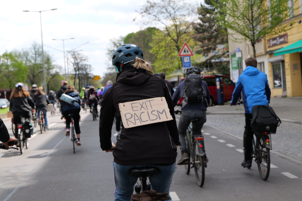 Eine Gruppe von Menschen, die auf Fahrrädern eine Straße entlangfahren, wobei eine Frau in der Mitte ein Schild hält, auf dem "Exit Racism" steht. Die Straße ist von Bäumen, Laternenpfählen und Gebäuden gesäumt und der Himmel ist im Hintergrund zu sehen.