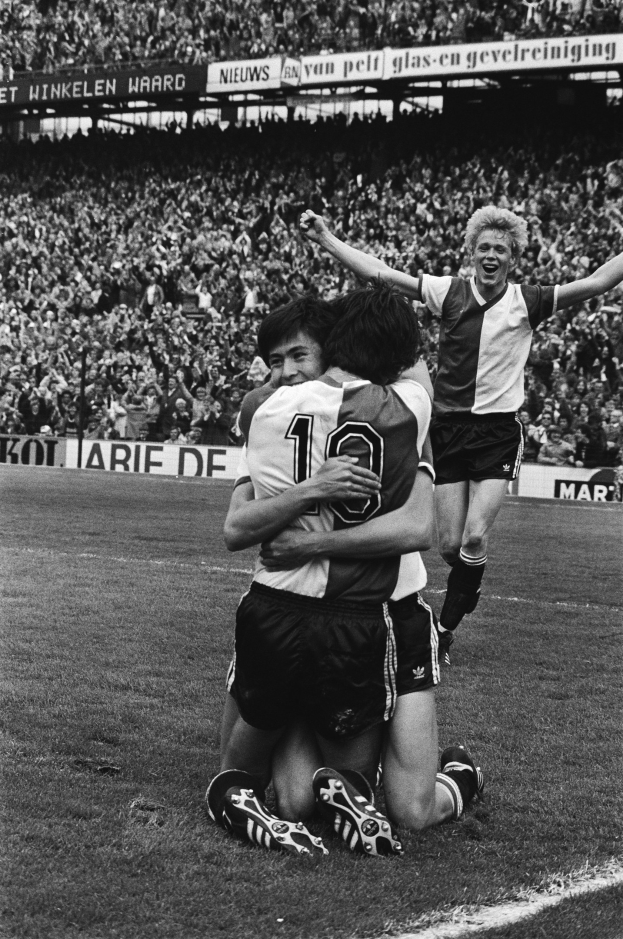 Drei Fußballspieler feiern ein Tor auf dem Boden, umgeben von einer jubelnden Menge in einer schwarz-weißen Stadionszene.