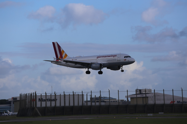 Germanwings Airbus A320-200 beim Abheben vom Frankfurter Flughafen, mit Gras darunter, einem Zaun im Vordergrund, Gebäuden und Bäumen im Hintergrund und einer bewölkten Himmel über