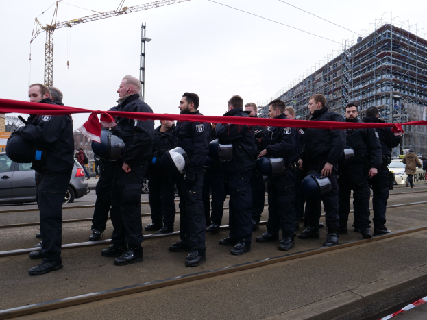 Eine Gruppe von Polizisten in schwarzen Uniformen und Helmen steht in einer Reihe auf einem Gleis mit einem roten Band davor, mit Fahrzeugen, Passanten, Gebäuden und einem klaren Himmel im Hintergrund.