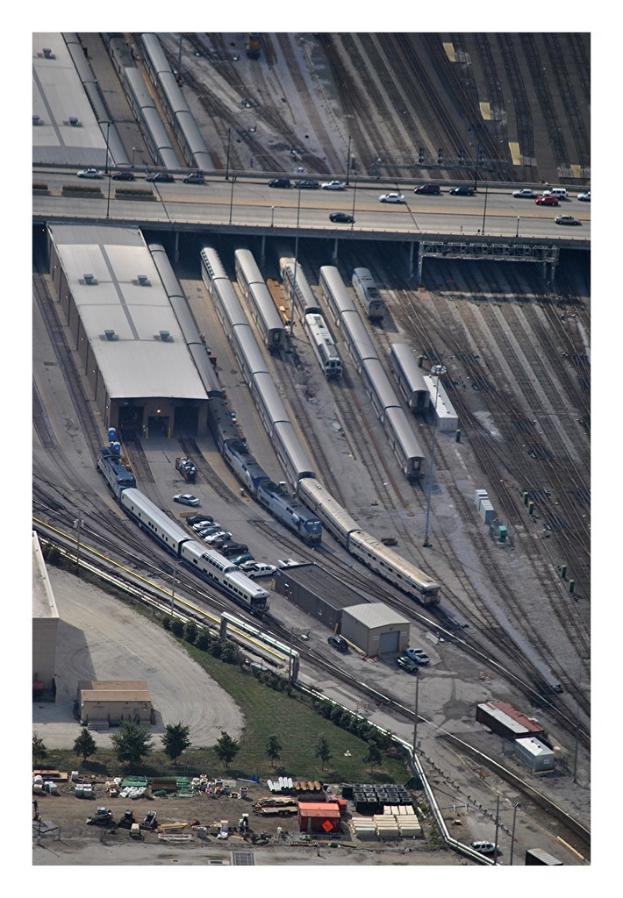Eine Brücke mit wenigen Fahrzeugen, eine darunter verlaufende Bahnstrecke mit wenigen Zügen und eine Landschaft mit Gebäuden, Bäumen, Masten und diversen Gegenständen und Fahrzeugen am Boden.